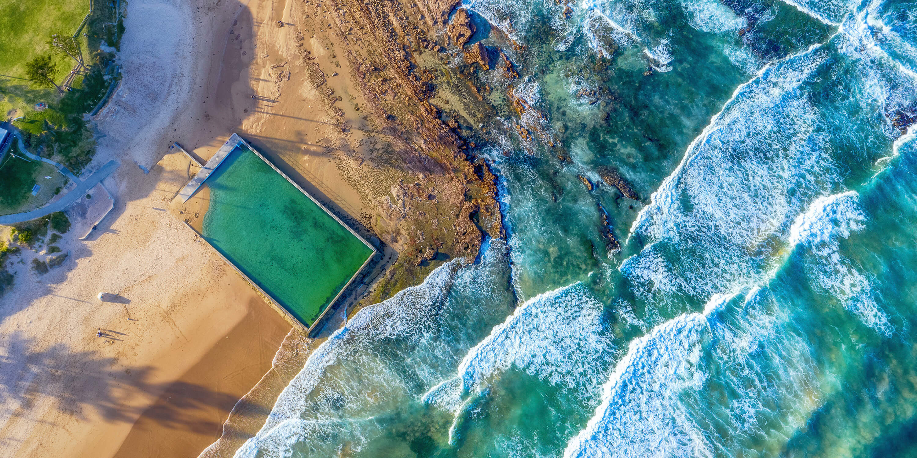 Aerial image of the ocean, a pool, the beach and grass.