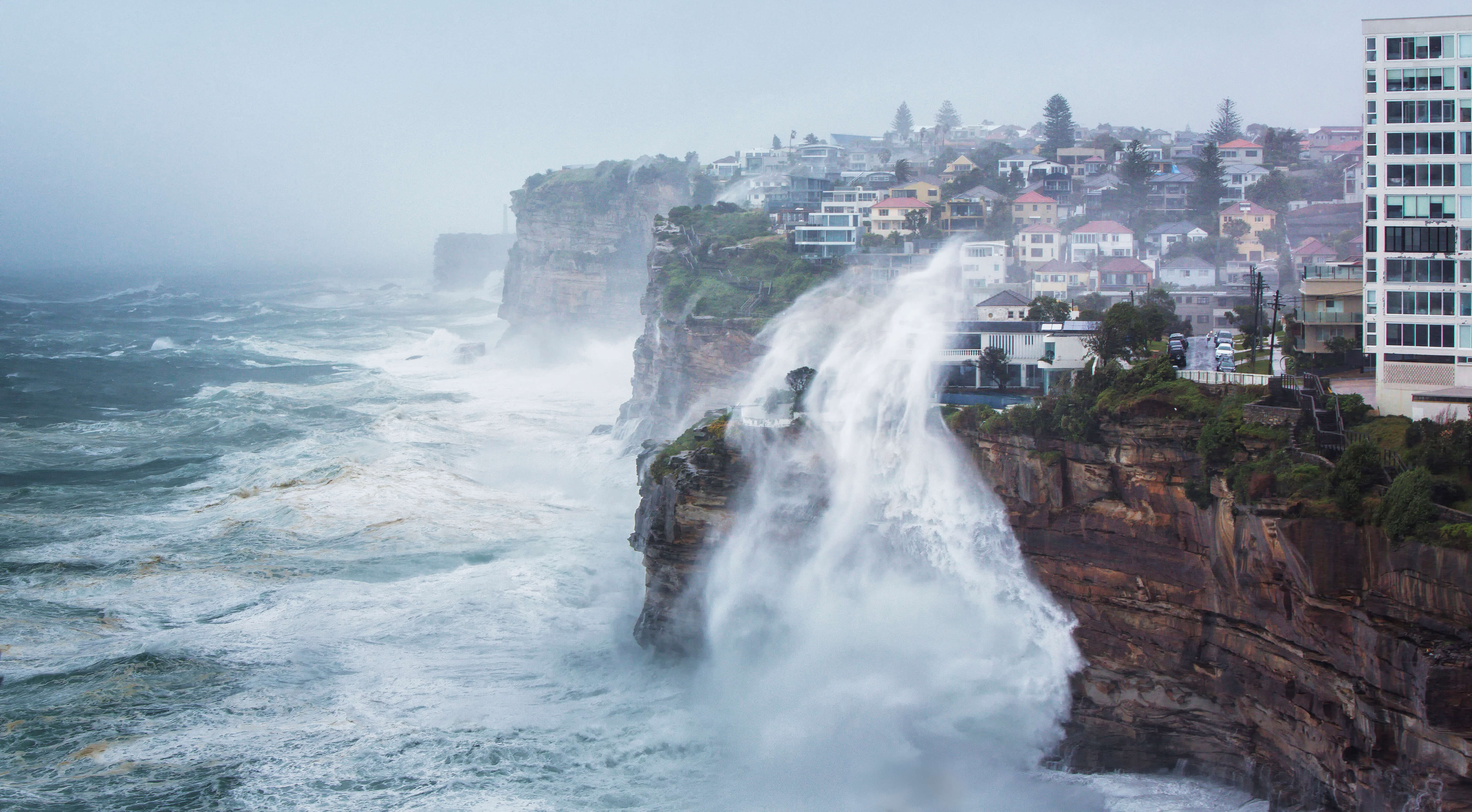 Coastline with habitations close to the edge of a cliff in a stormy weather with an impressive wave splashing up the cliff to the houses